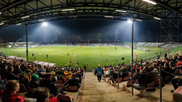 Estádio de futebol à noite com os projetores acesos sobre o relvado e adeptos nas bancadas